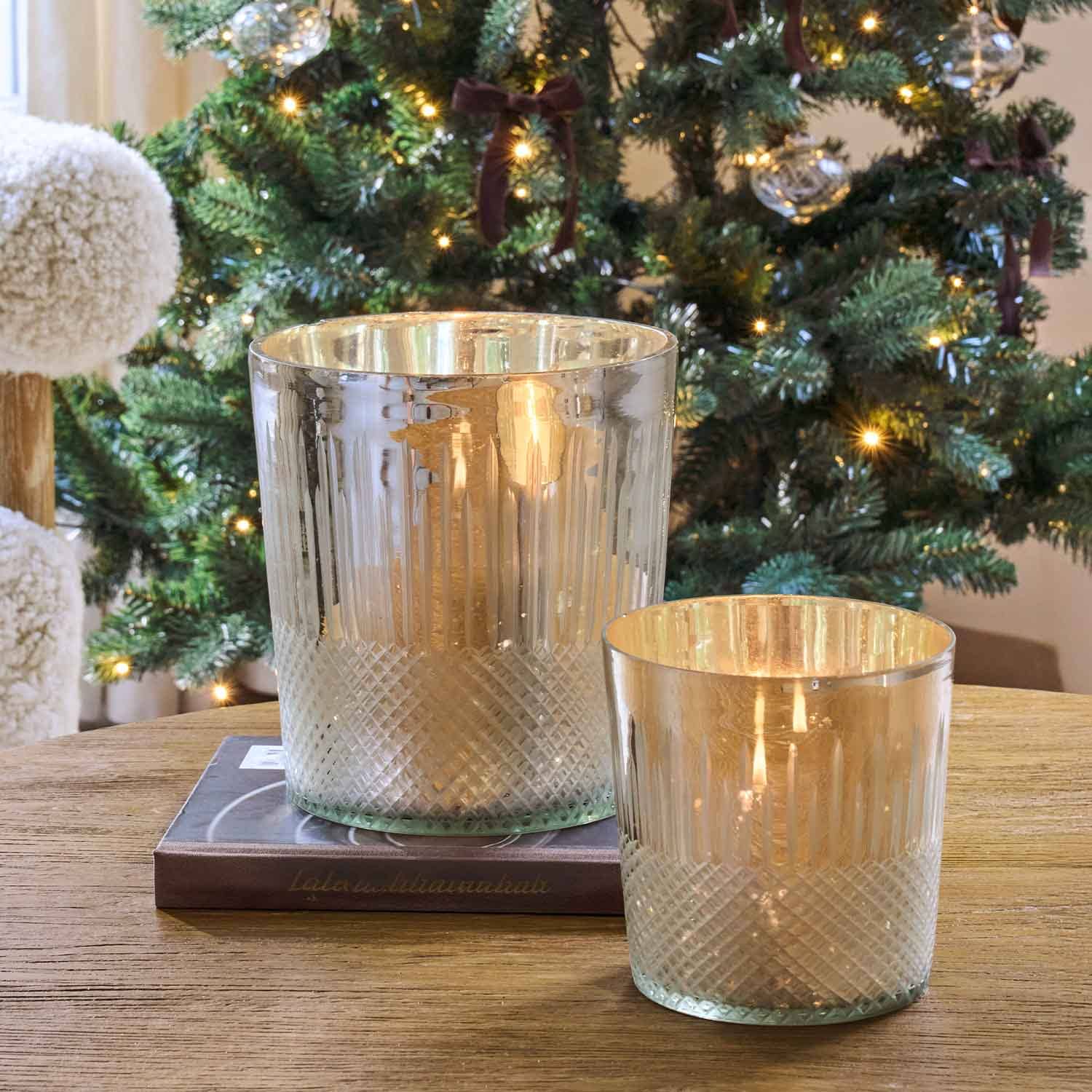 Zwei Glas-Teelichthalter mit metallischem Glanz auf Holztisch vor festlich geschmücktem Weihnachtsbaum