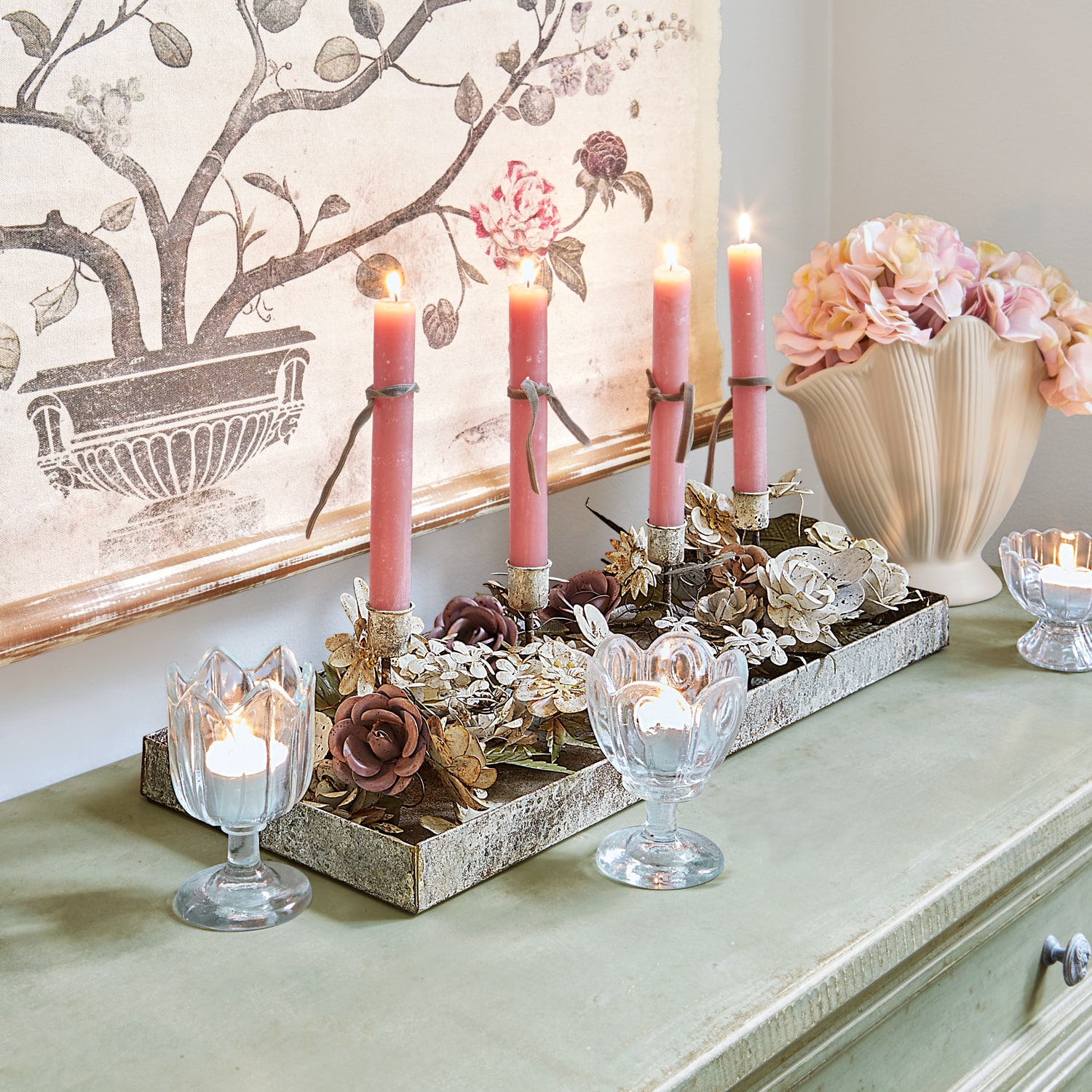 Dekorativer Kerzenhalter mit Metallblumen und rosa Kerzen auf grünem Sideboard mit Vase