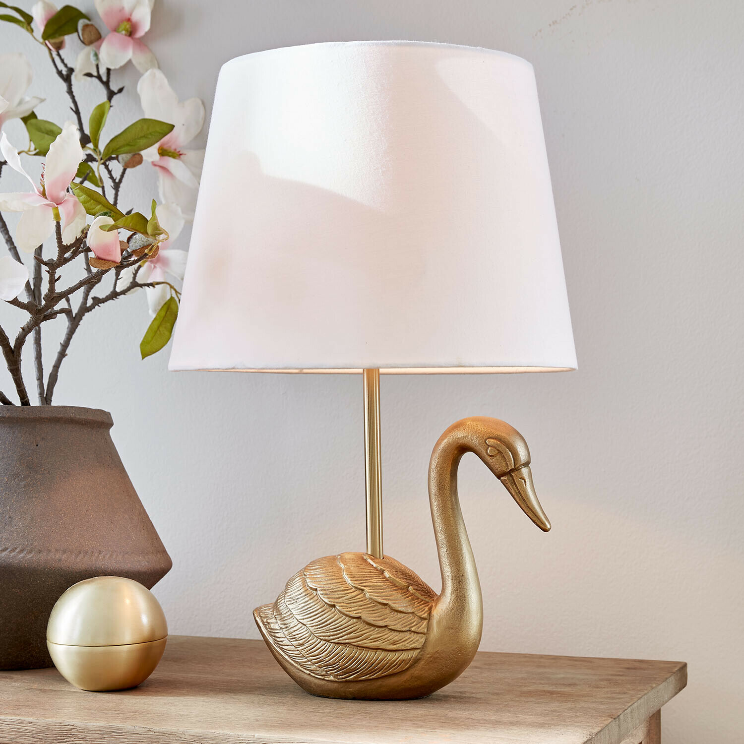 Goldene Tischlampe in Schwanform mit weißem Lampenschirm auf Holztisch mit Blumenarrangement
