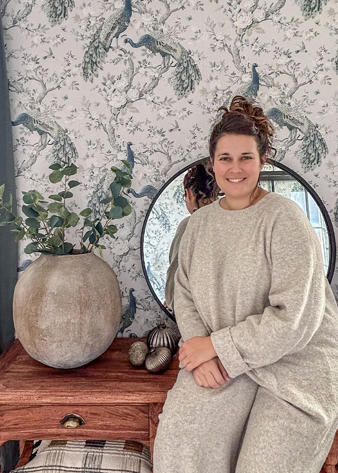 Frau im grauen Pullover sitzt an einem Holztisch mit großer Keramikvase und Spiegel vor einer Pfauen-Tapete