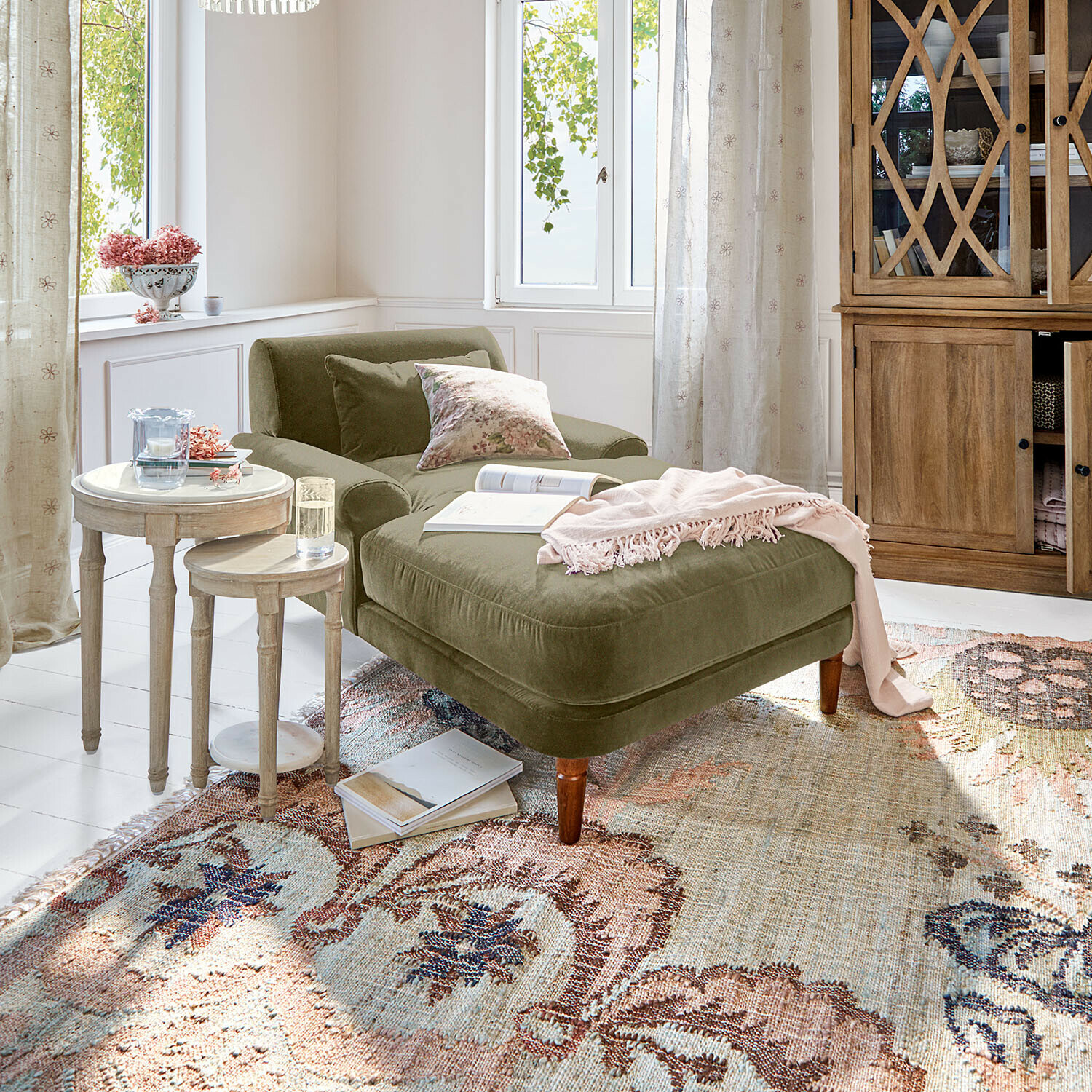 Chaiselounge Jouvrelle Grüne Samt-Chaiselongue mit Kissen und rosa Decke in hellem Wohnzimmer mit Holzmöbeln