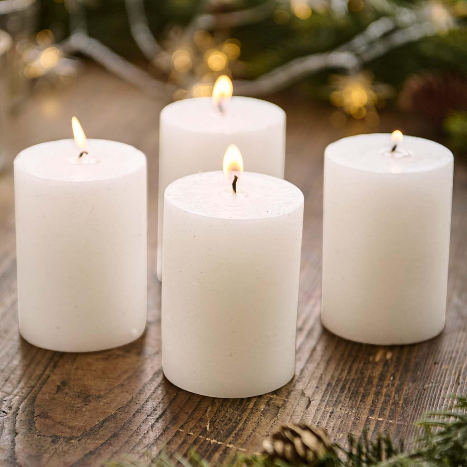 Vier weiße Stumpenkerzen auf Holztisch mit weihnachtlicher Dekoration im Hintergrund