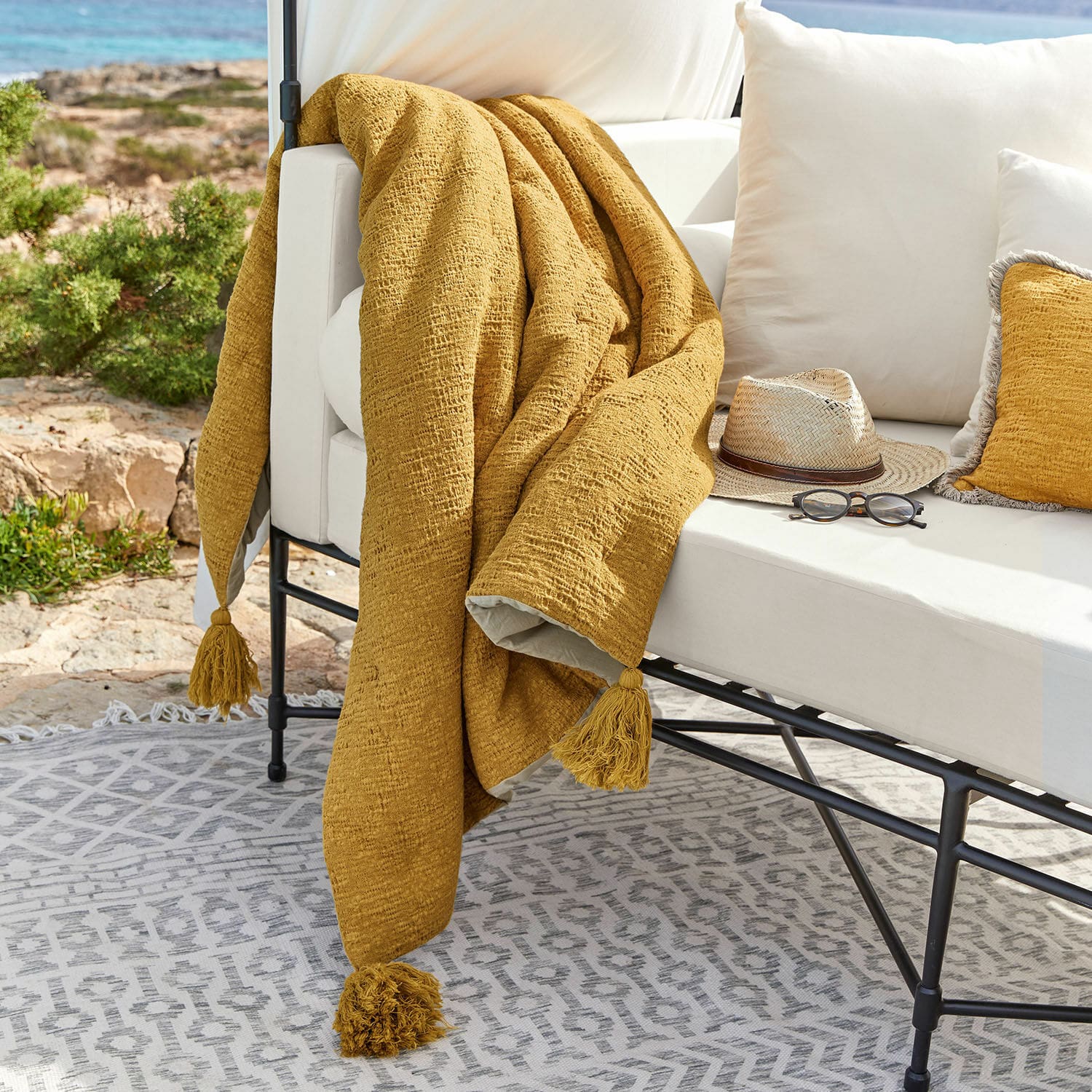Plaid Rabea Gelbe Tagesdecke mit Quasten auf weißem Outdoor-Sofa, Terrasse mit Sonnenbrille und Hut dekoriert