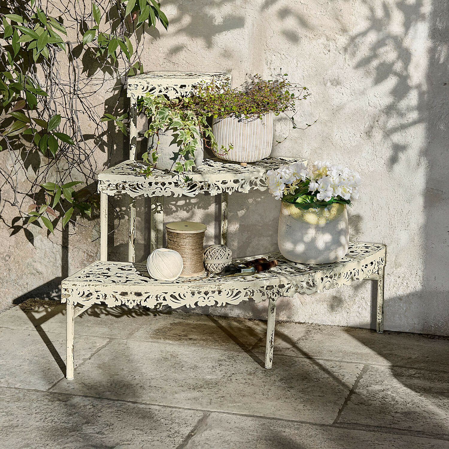 Pflanztreppe Cresselay Cremefarbene Metall-Etagere mit filigranem Muster, bepflanzt und mit Gartengeräten auf Steinboden