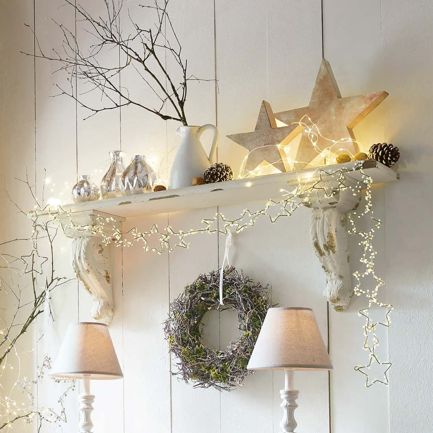 Weiße Holzregal-Dekoration mit sternförmiger Lichterkette, Kranz und Tischlampen im Landhausstil