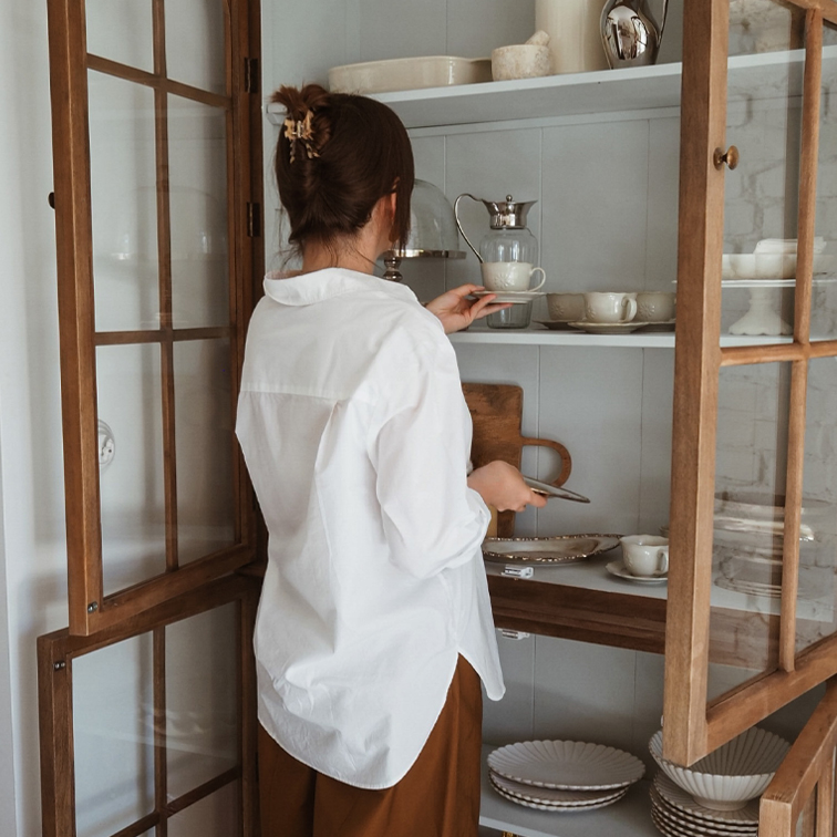 Frau in weißem Hemd nimmt Porzellantasse aus Holzvitrine mit Geschirr im Landhausstil