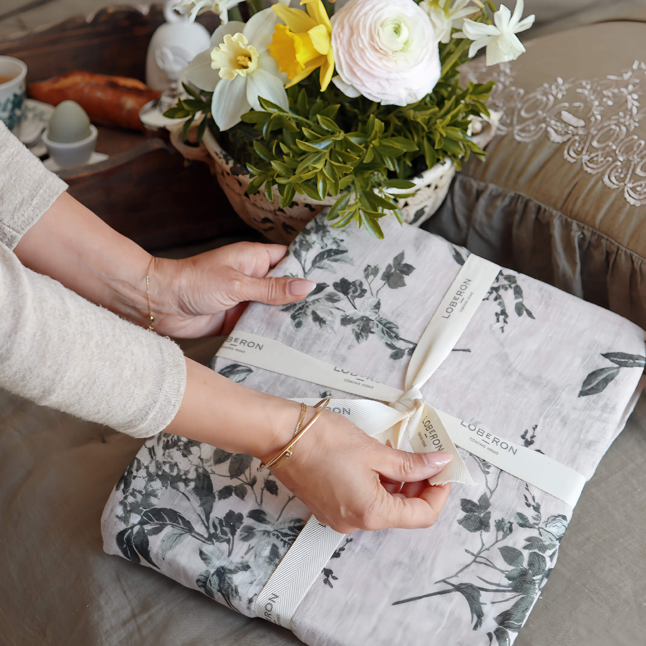 Hände öffnen graue Leinenbettwäsche mit Pflanzenmuster, Blumenarrangement im Hintergrund