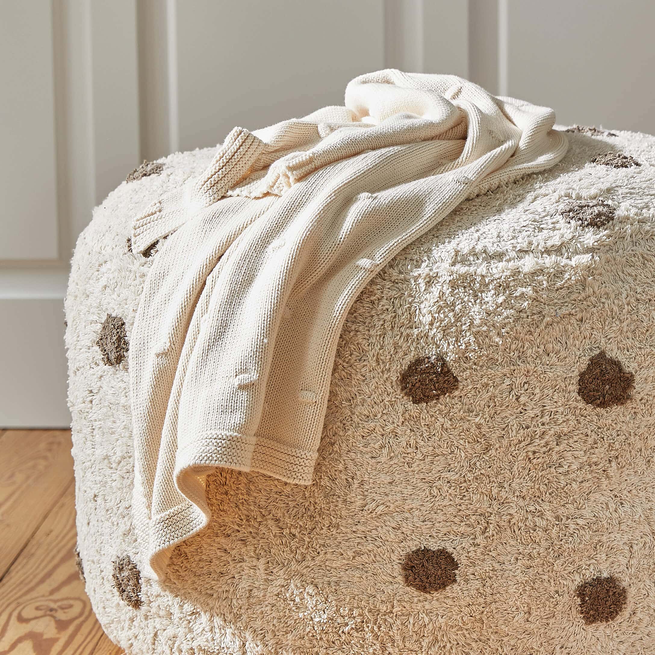 Beiger Strickpullover auf cremefarbenem Pouf mit braunen Punkten, weiches Material, Wohnaccessoire