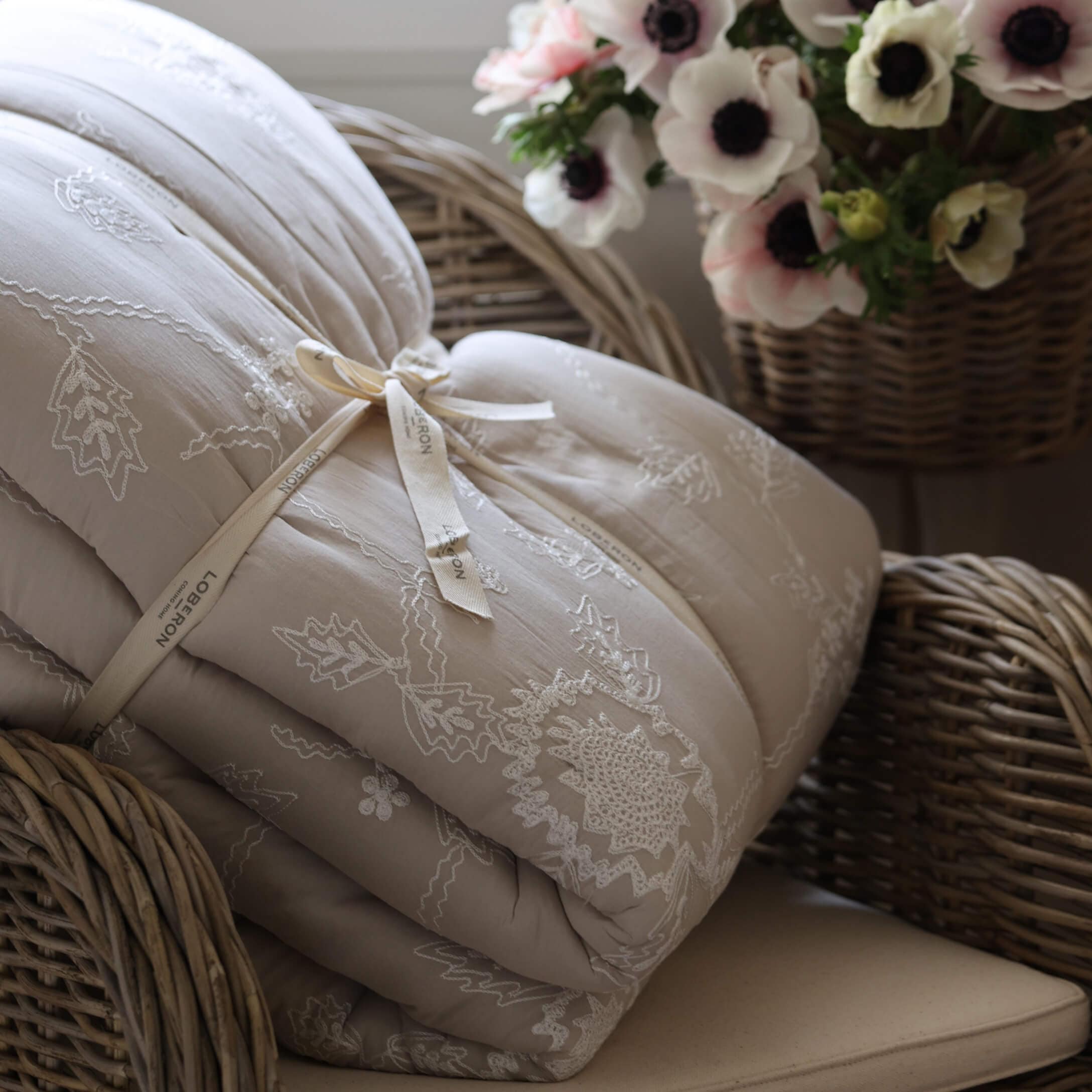 Beige gesteppte Tagesdecke mit Stickerei auf einem Korbsessel neben einem Blumenkorb, Wohnaccessoire