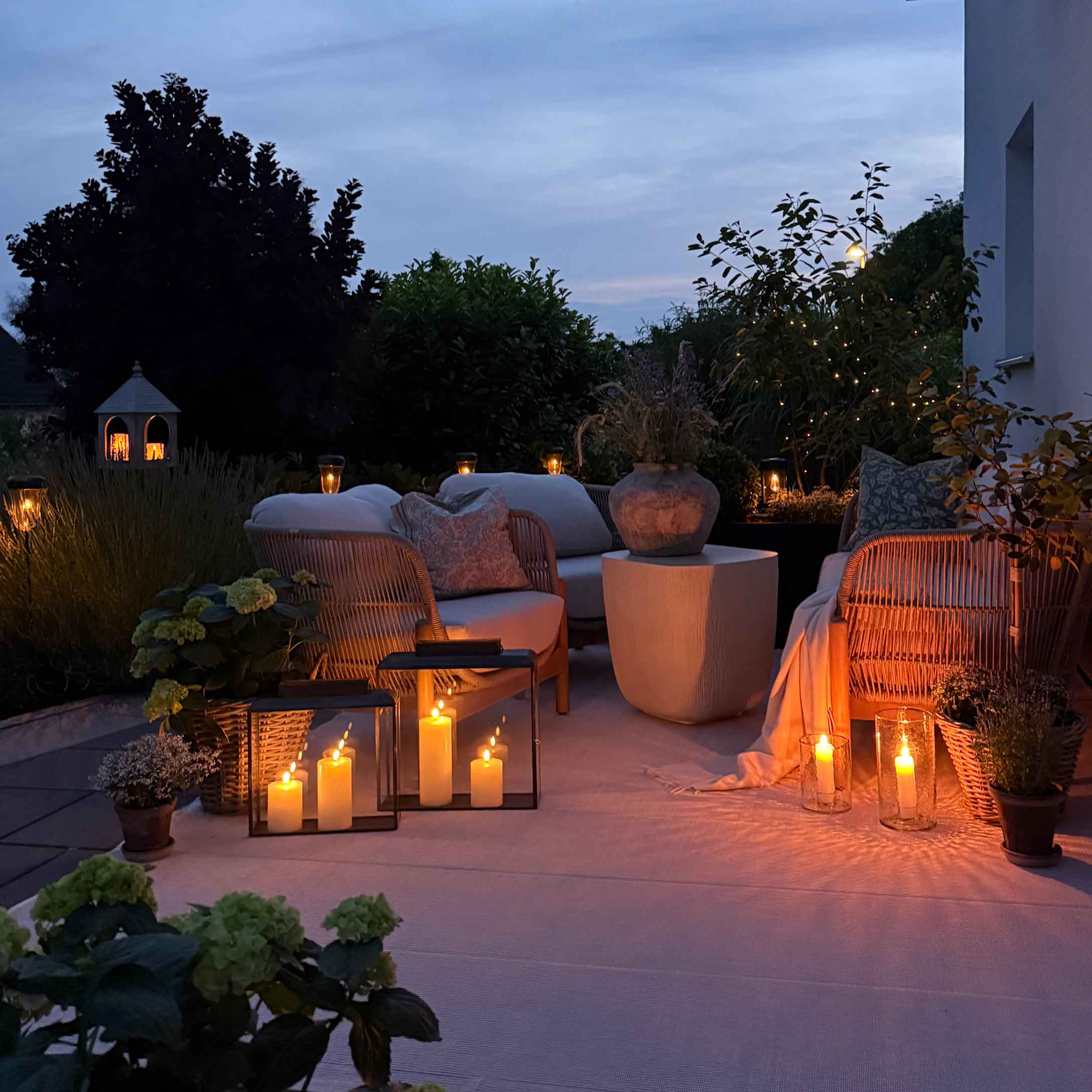 Gemütlicher Garten mit weißen Kissen auf Möbeln und dekorativen Kerzen am Abend