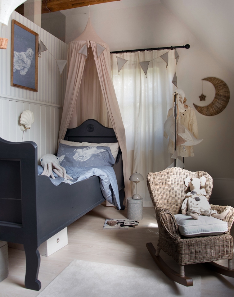 Kinderzimmer mit schwarzem Holzbett, beigem Baldachin und Rattanschaukelstuhl