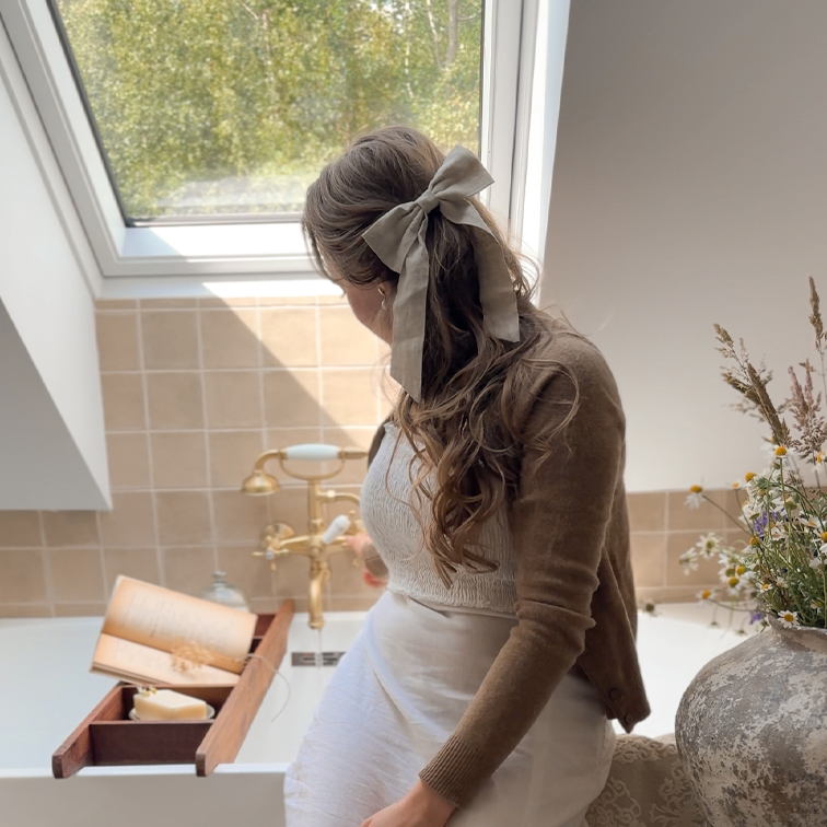 Frau mit beiger Leinenhaarschleife sitzt am Bad mit Holzablage und Buch, natürliches Licht durch Dachfenster