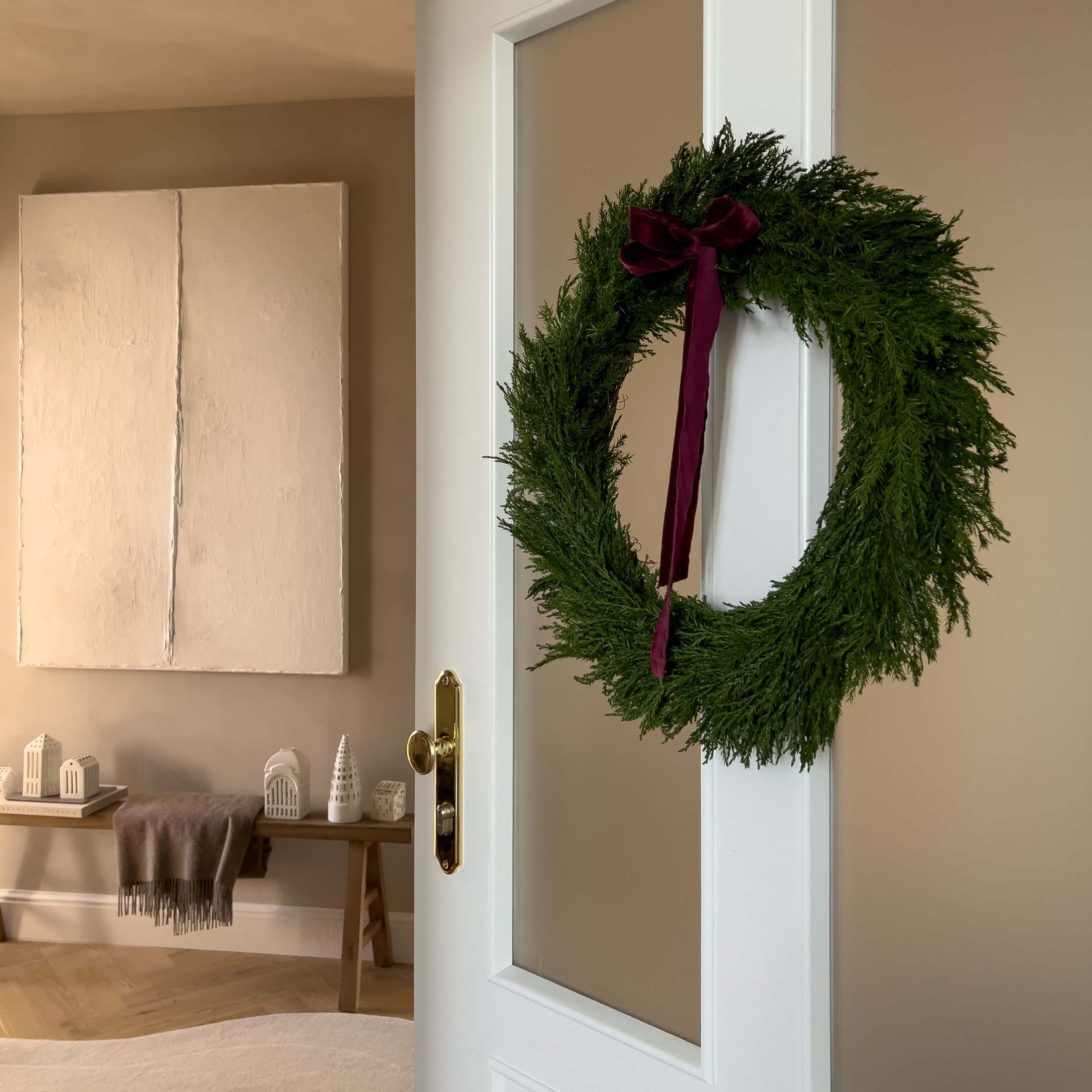 Grüner Weihnachtskranz mit bordeauxroter Schleife an weißer Tür in gemütlichem Raum