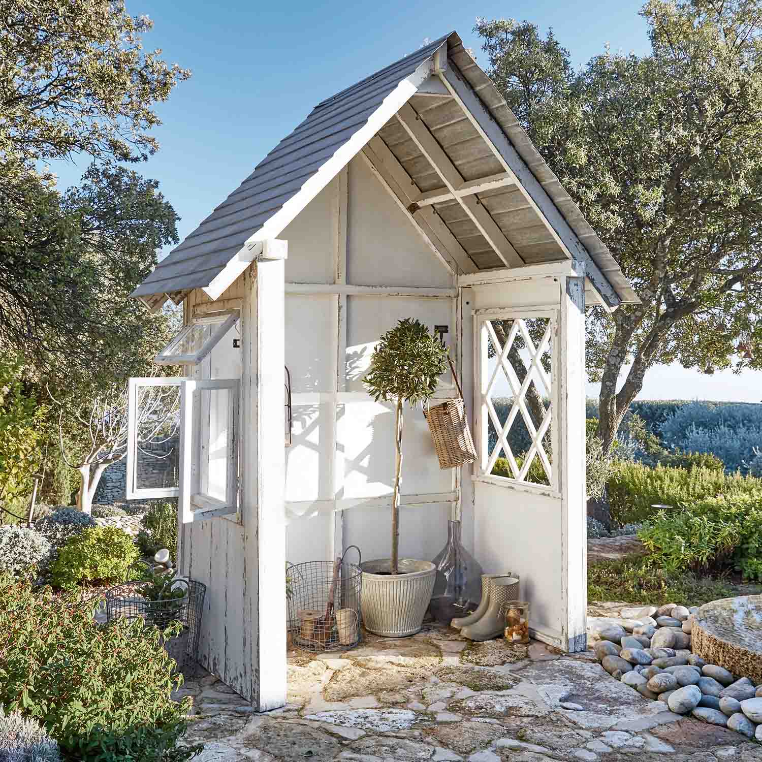 Weißes Holz-Gartenhaus mit Fenstern und Pflanzen auf Steinboden in sonnigem Garten