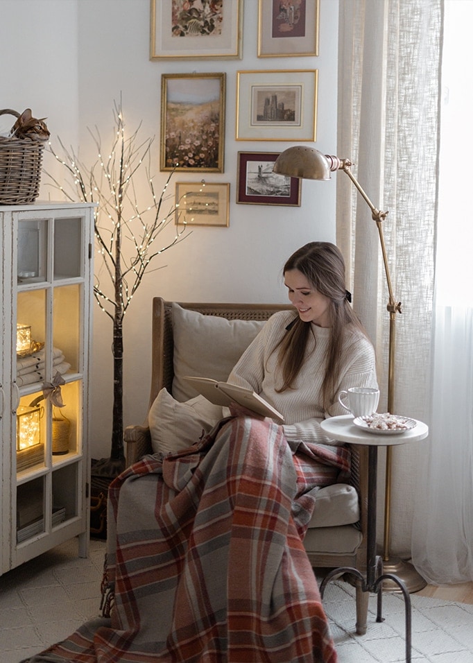 Frau liest Buch unter warmem Plaid in gemütlichem Wohnzimmer mit Stehlampe und Wandbildern