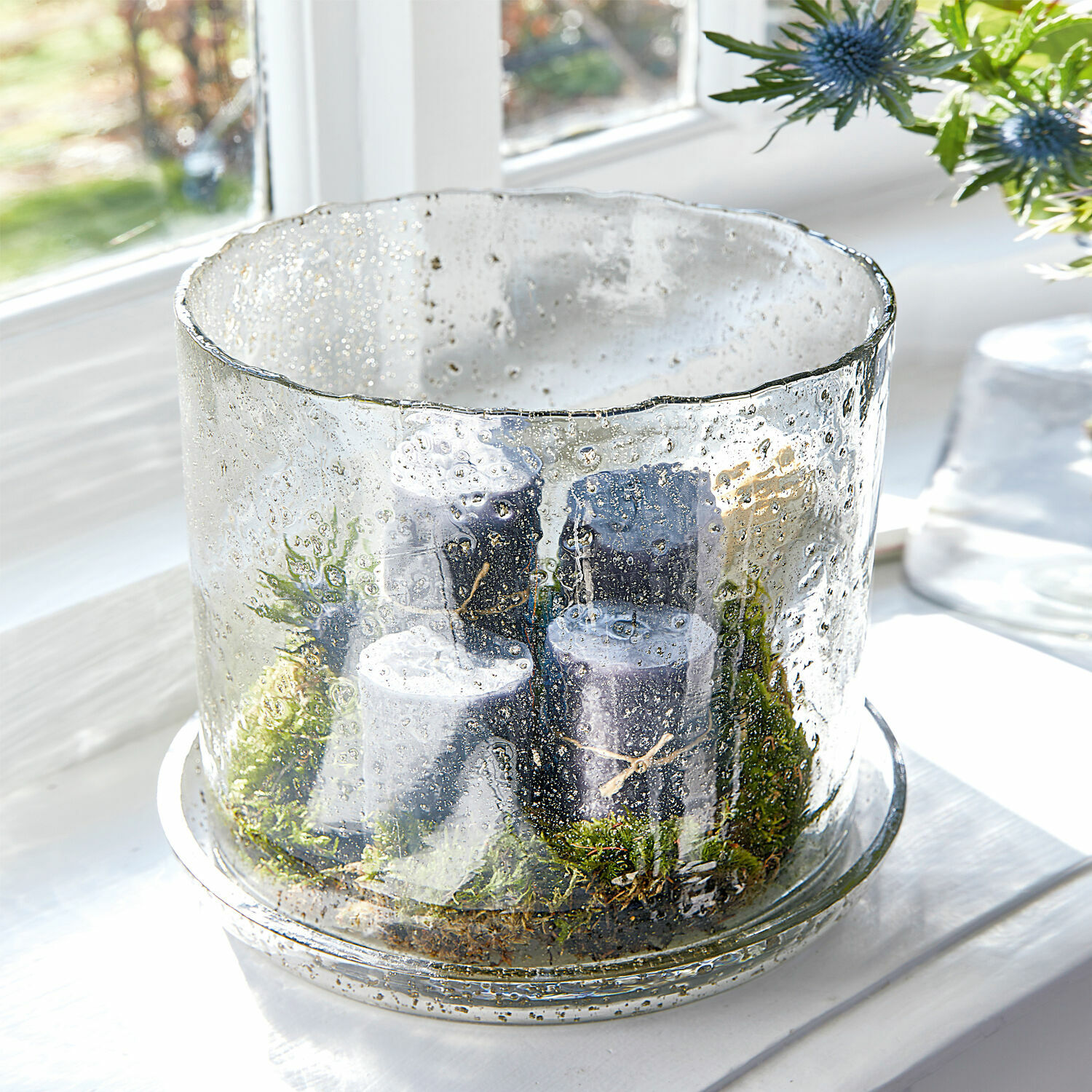 Glaswindlicht mit Luftblasen und schwarzen Kerzen auf grünem Moos auf Fensterbank