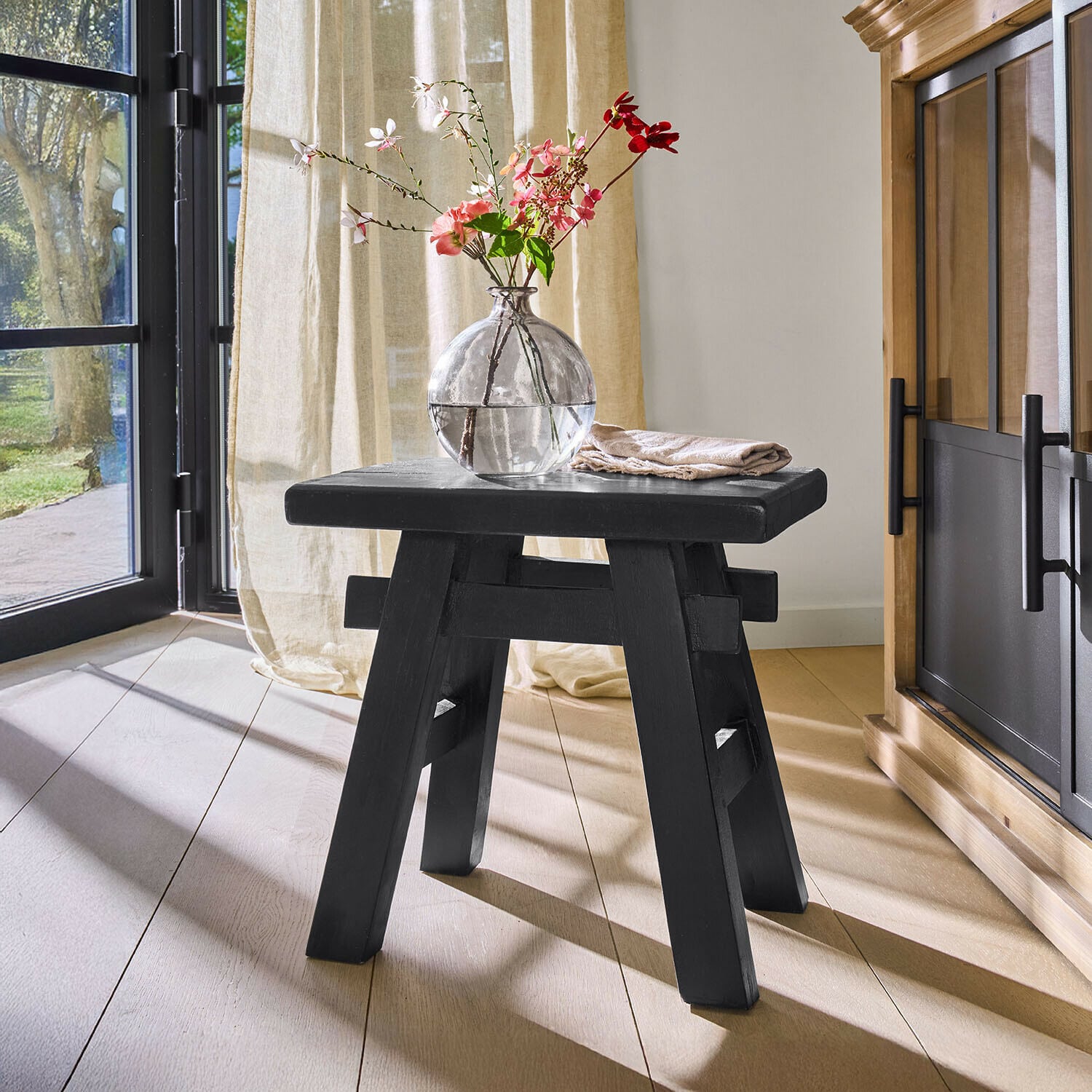Hocker Oaklynne Schwarzer Holzschemel mit einfacher Konstruktion in hellem Raum mit Vase und Blumen