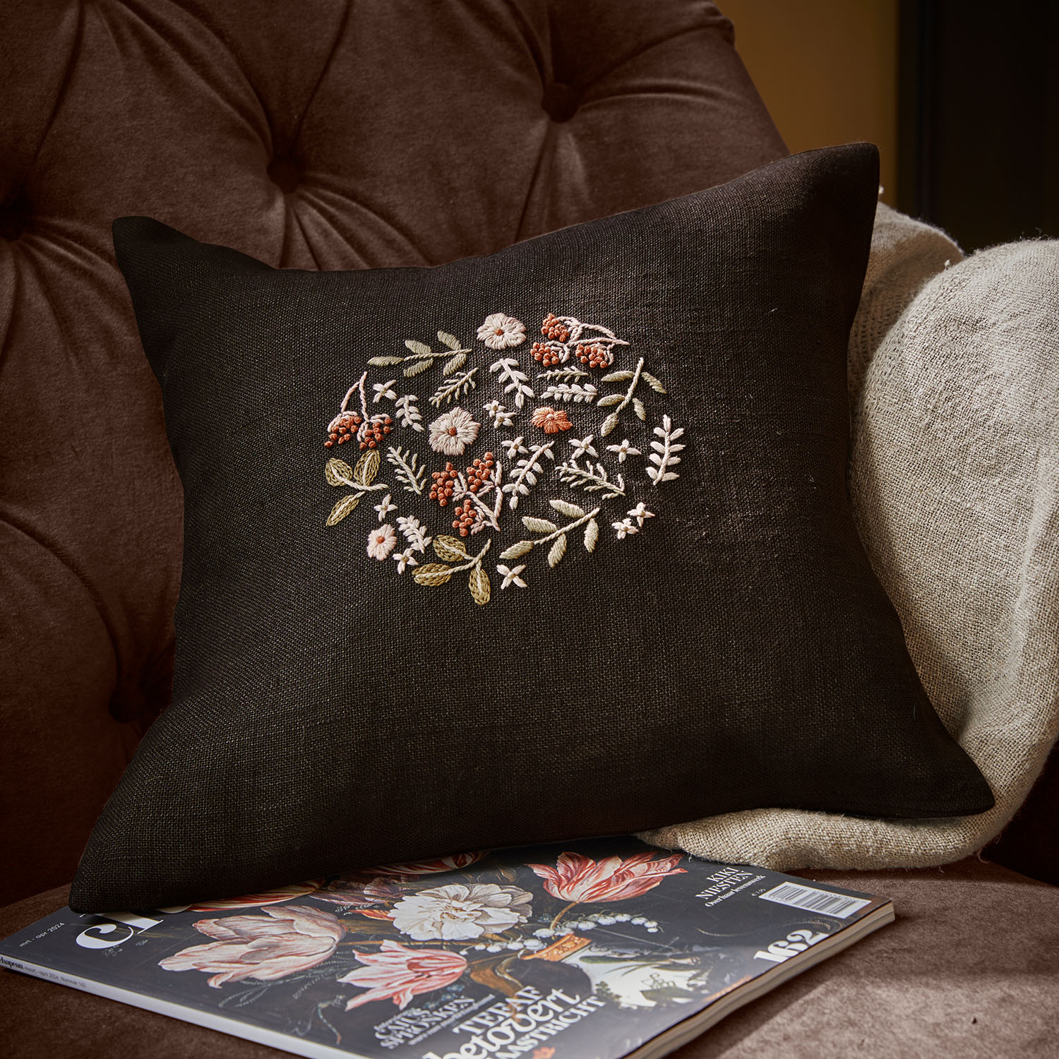 Schwarzes Leinenkissen mit pastellfarbenem Blumenstickmuster auf braunem Sessel