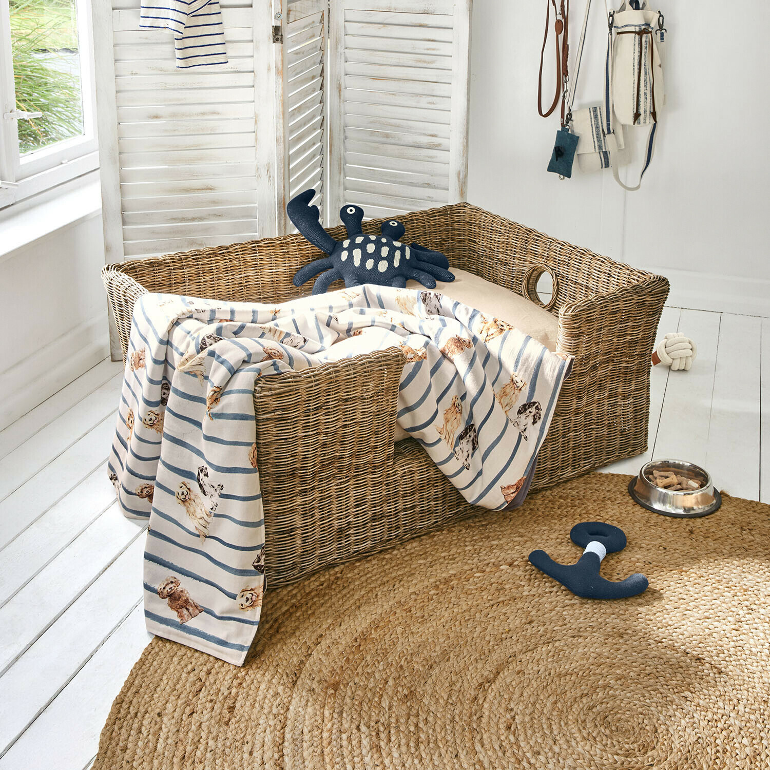 Wicker-Hundebett mit beigem Kissen, gestreifter Decke mit Hunde-Motiv, Spielzeug und Futternapf mit Leckerlis