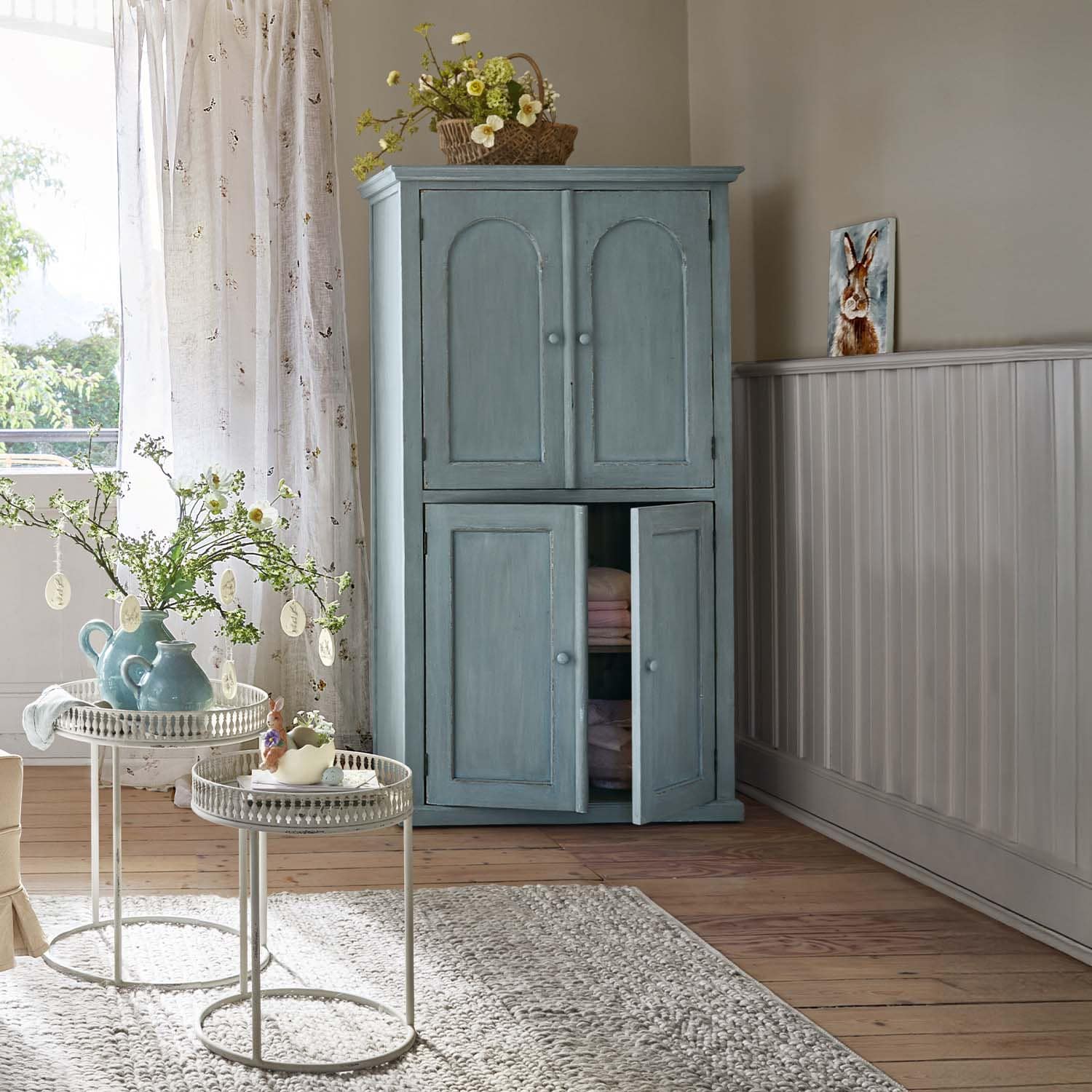 Hellblauer Holzschrank mit zwei Türen in gemütlichem Raum mit Blumenarrangements und Teppich