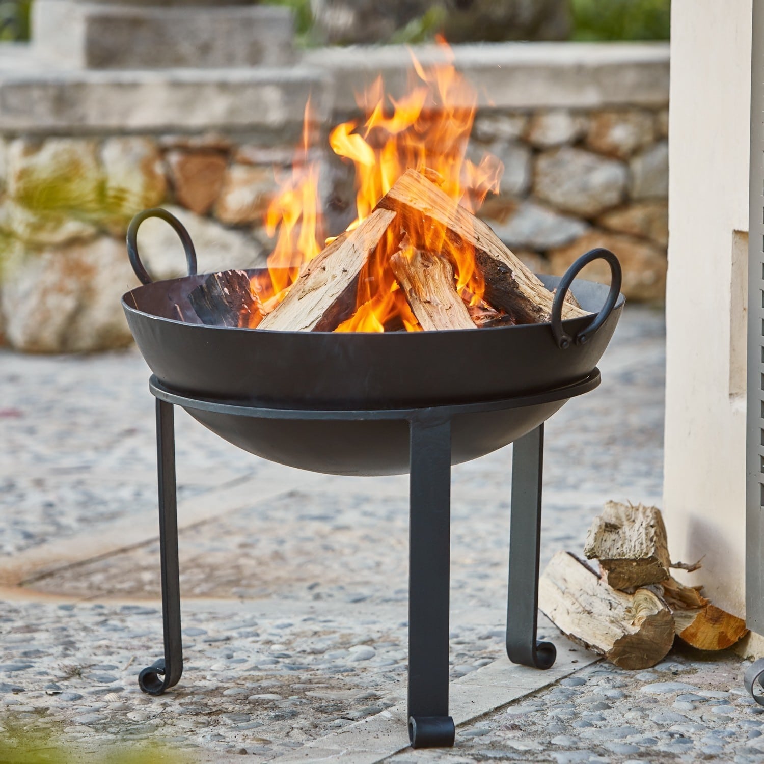Feuerschale Harrogate Schwarzes Metall-Feuerkorb mit brennendem Holz auf steiniger Terrasse im Garten