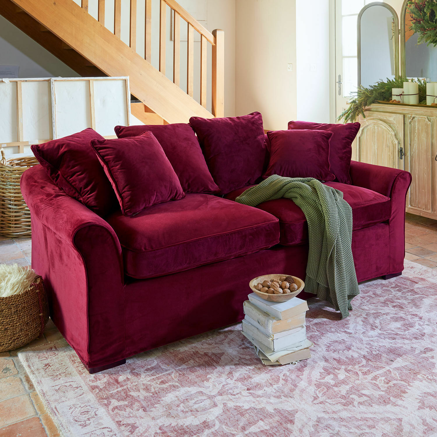 Bordeauxrotes Samtsofa mit Kissen und grünem Strickplaid auf rustikalem Teppich im Wohnzimmer