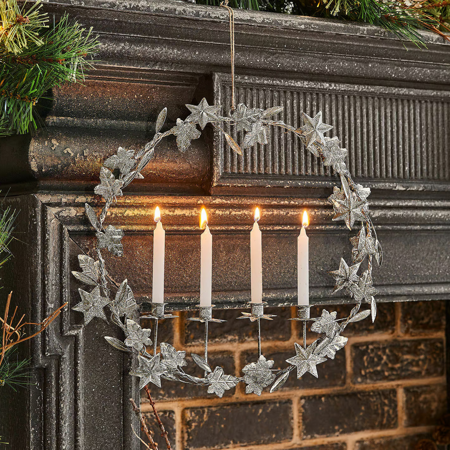Metallkranz mit Blättern und vier weißen brennenden Kerzen an einem Kamin, Weihnachtsdeko