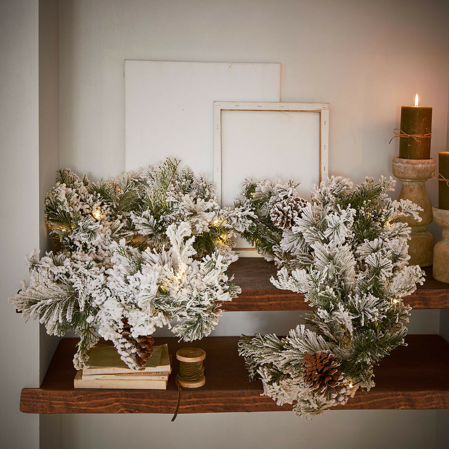 Weihnachtlicher Kranz aus grünen, schneebedeckten Zweigen mit Zapfen und LED-Lichterkette auf Holzregal