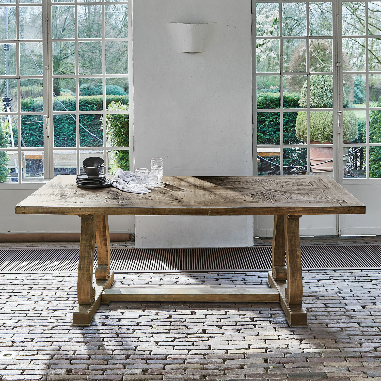 Esstisch aus Naturholz mit dekorativen Beinen vor großen Fenstern und Ziegelboden im Raum