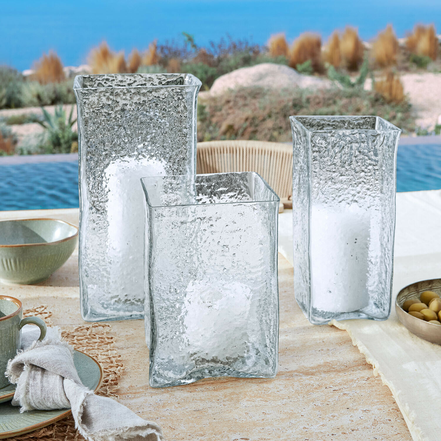 Windlicht 3er Set Xarlac Drei durchsichtige, strukturierte Glaslaternen mit weißen Kerzen auf einem Tisch am Pool
