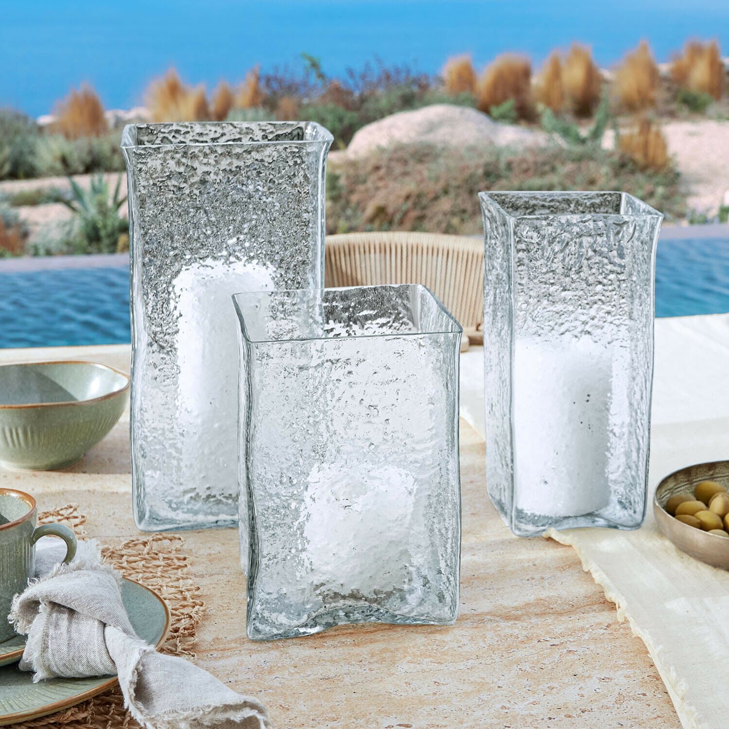 Windlicht 3er Set Xarlac Drei durchsichtige, strukturierte Glaslaternen mit weißen Kerzen auf einem Tisch am Pool