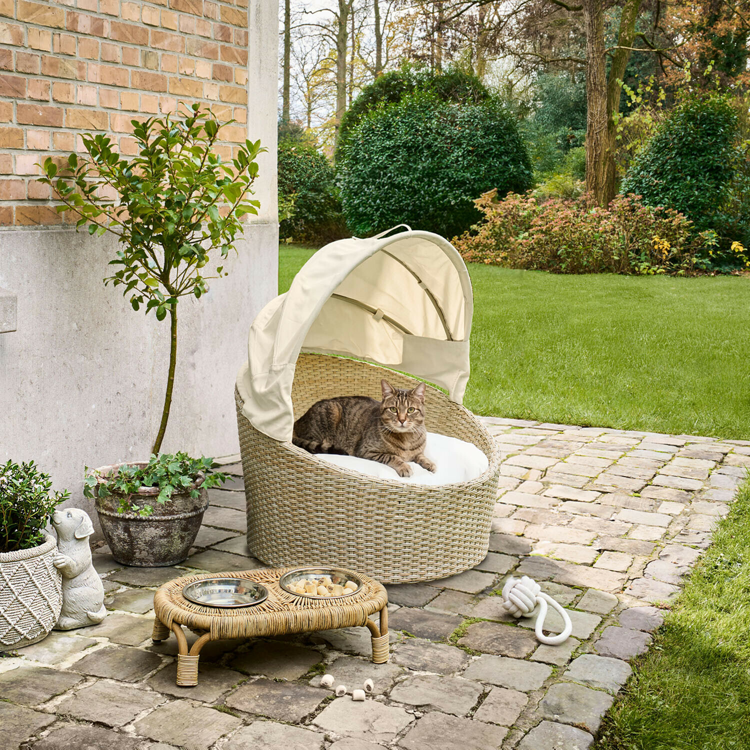 In- & Outdoor-Haustierkorb Brinelza Ovales Katzenbett aus beigem Rattan mit weißem Kissen und Dach, auf gepflasterter Terrasse im Garten