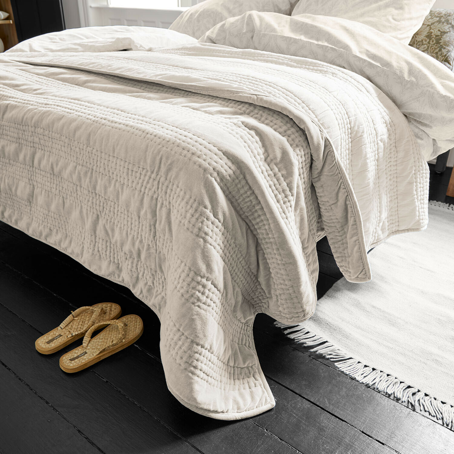 Beige gesteppte Tagesdecke aus weichem Material auf Bett mit heller Bettwäsche und Hausschuhen daneben