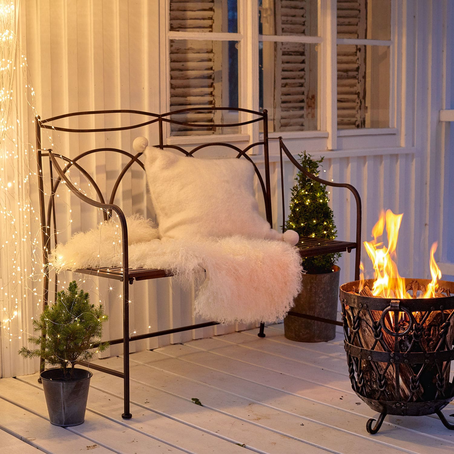 Bank Jablines Metallbank mit weißem Kissen und Fellüberwurf neben Feuerkorb auf Terrasse mit Lichterketten-Dekor