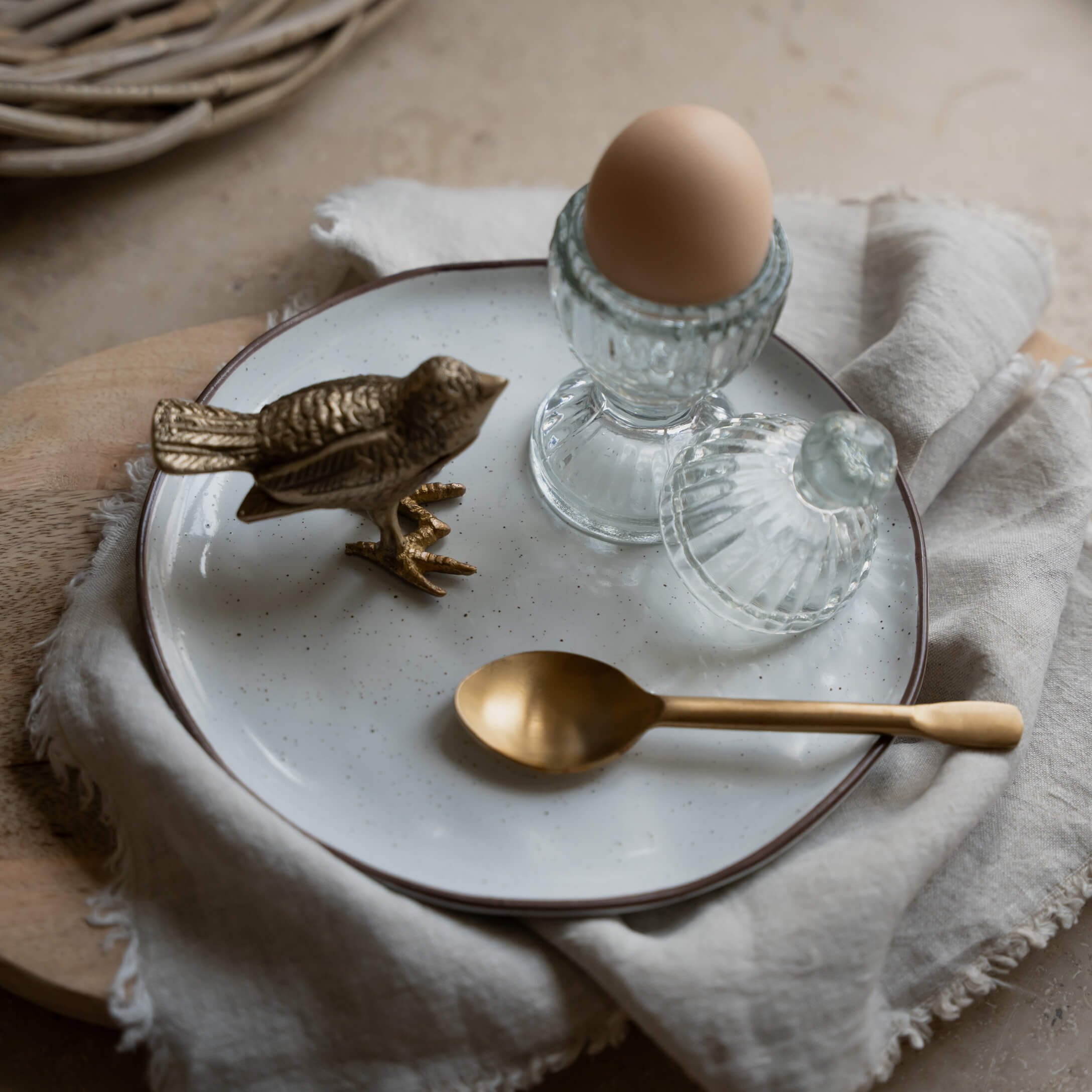 Glas-Eierbecher mit Deckel und braunem Ei auf weißem Teller mit Messinglöffel und Vogel-Figur