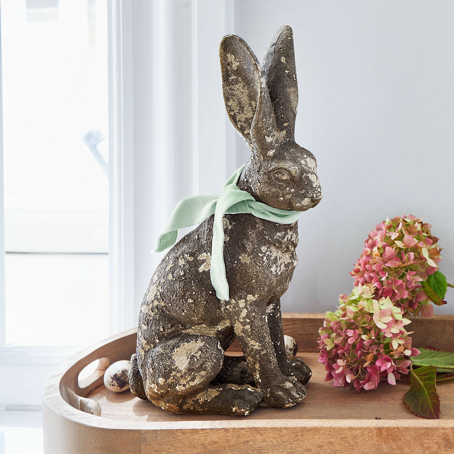 Dekorativer Hase aus patiniertem Stein mit grünem Band auf Holztablett mit Blumen