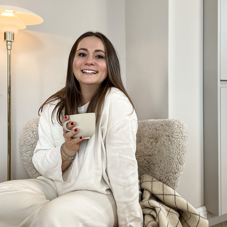 Lächende Frau in weißem weichem Outfit sitzt mit Tasse auf gemütlichem Sessel in warmem Raum