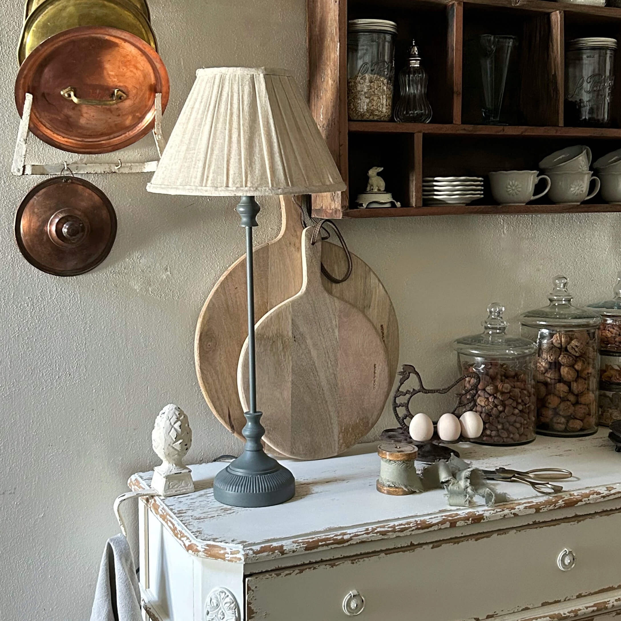 Tischlampe mit grauem Sockel und cremefarbenem Lampenschirm auf weißem, antik wirkendem Schrank in rustikaler Küche