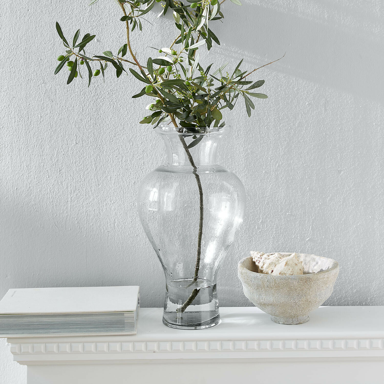 Vase Cantellia Transparente Glasvase mit grünen Zweigen auf weißem Regal mit Buch und Muschelschale