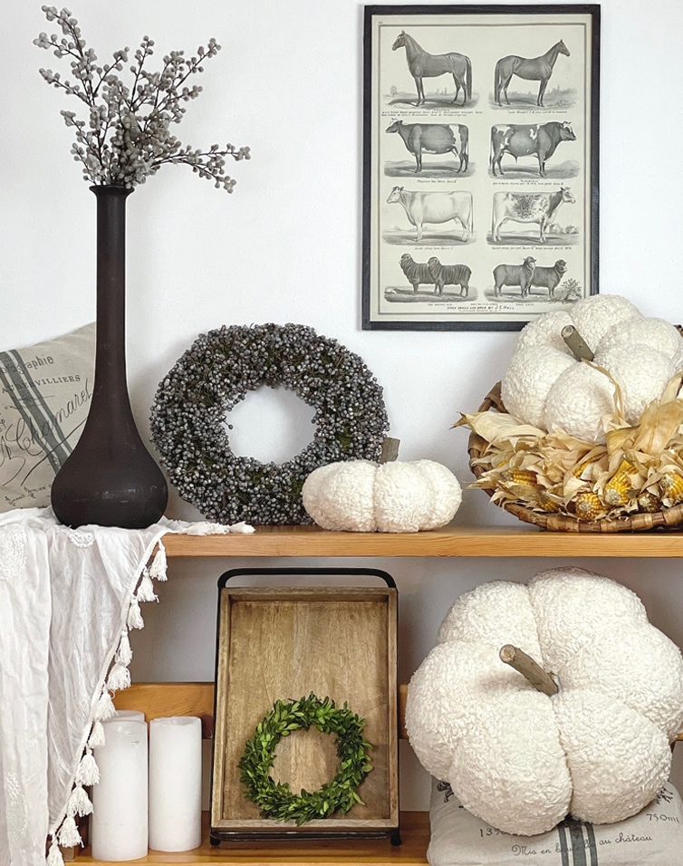 Herbstliche Dekoration mit weißen Plüsch-Kürbissen, grünen Kränzen und schwarzer Vase auf Holzregal
