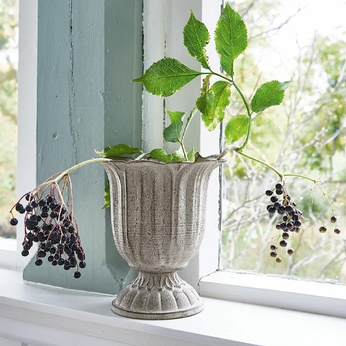 Graue Keramikvase mit grünen Blättern und schwarzen Beeren auf Fensterbank dekoriert