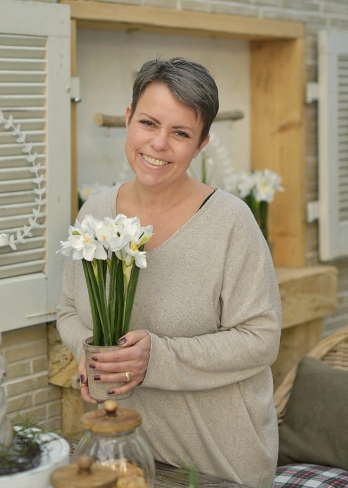 Frau hält Topf mit weißen Blumen in natürlicher Osterdekoration, trägt beigen Pullover