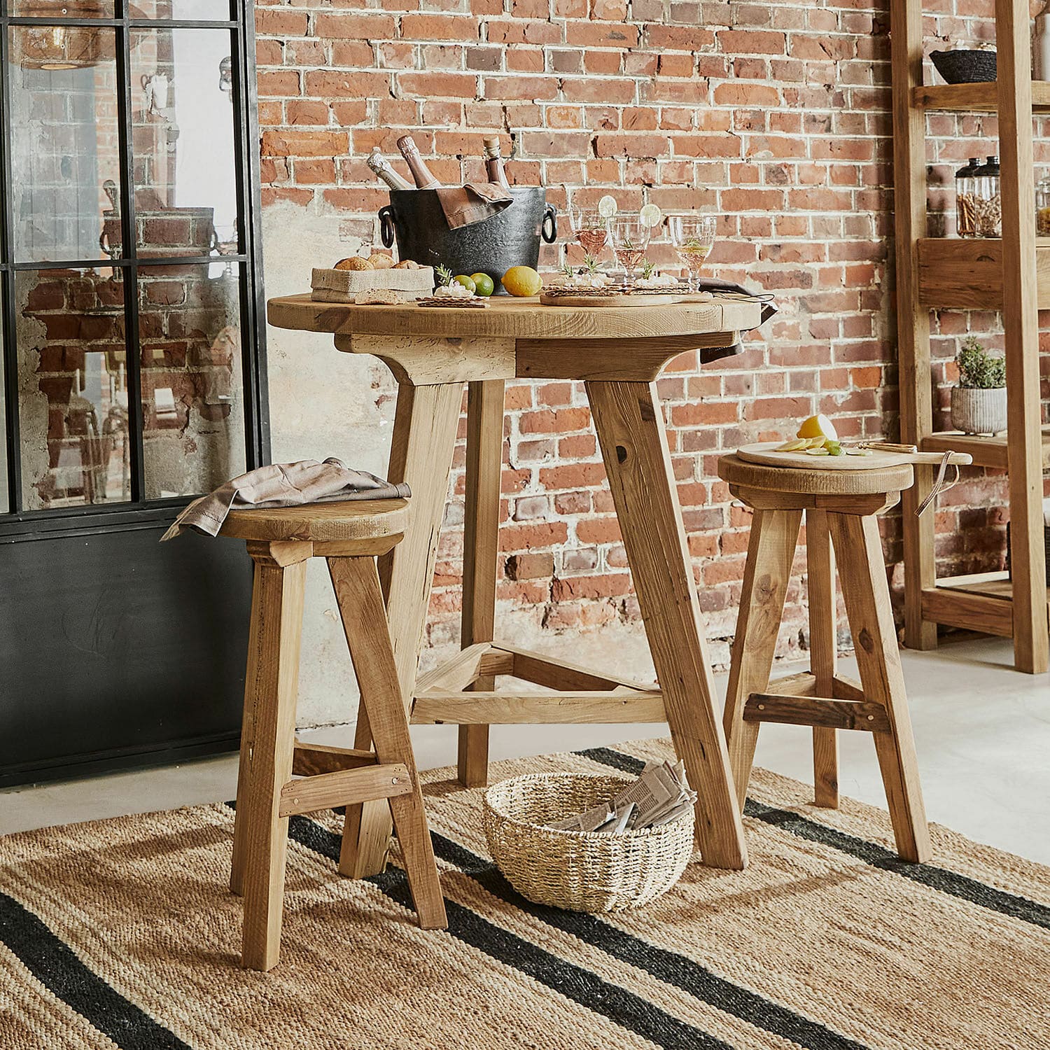 Sitzgruppe Stonington Hoher Holztisch mit zwei Hockern in Naturholz auf Jute-Teppich vor Backsteinwand im Industrie-Stil