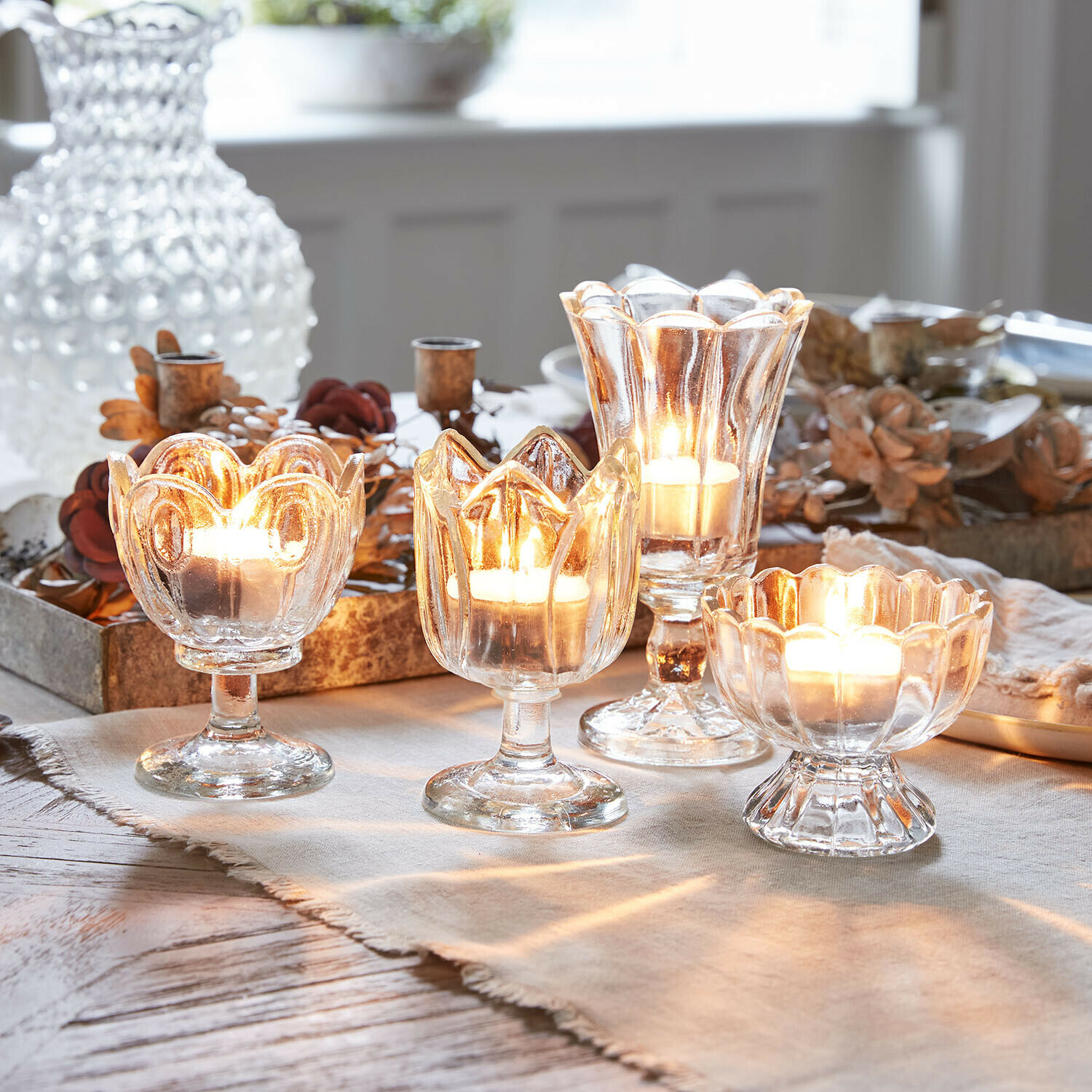 Vier transparente Glas-Teelichthalter auf Fuß mit brennenden Kerzen, Tischdekoration