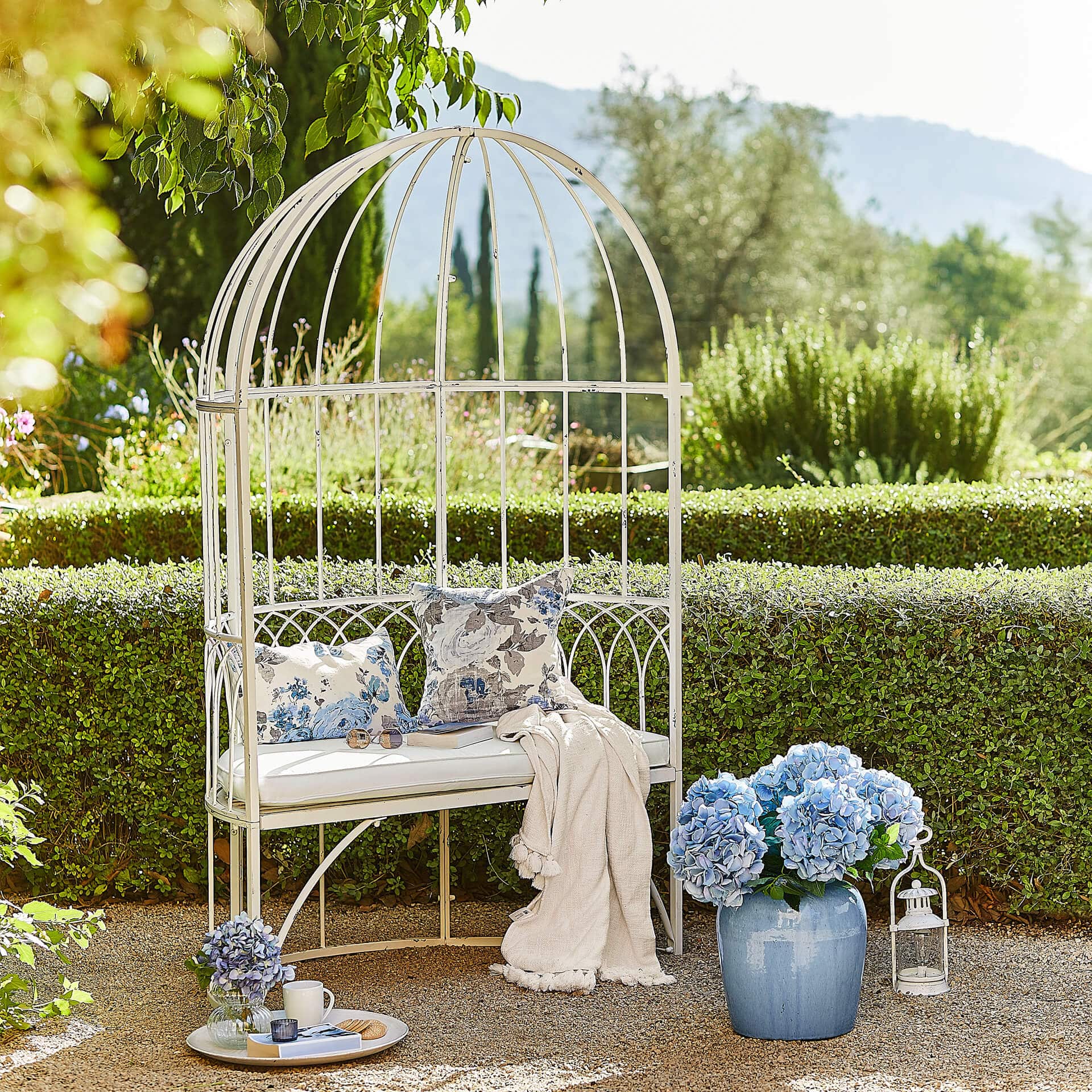 Weiße Metall-Gartenbank mit Kissen und blauen Blumen in Vase vor grüner Hecke
