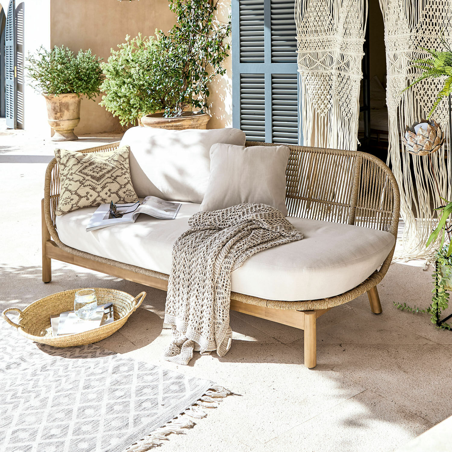 Chaiselongue FSC®-100% Belvidere Rattan- und Holzsofa mit cremefarbenen Kissen und Decke auf heller Terrasse mit Pflanzen