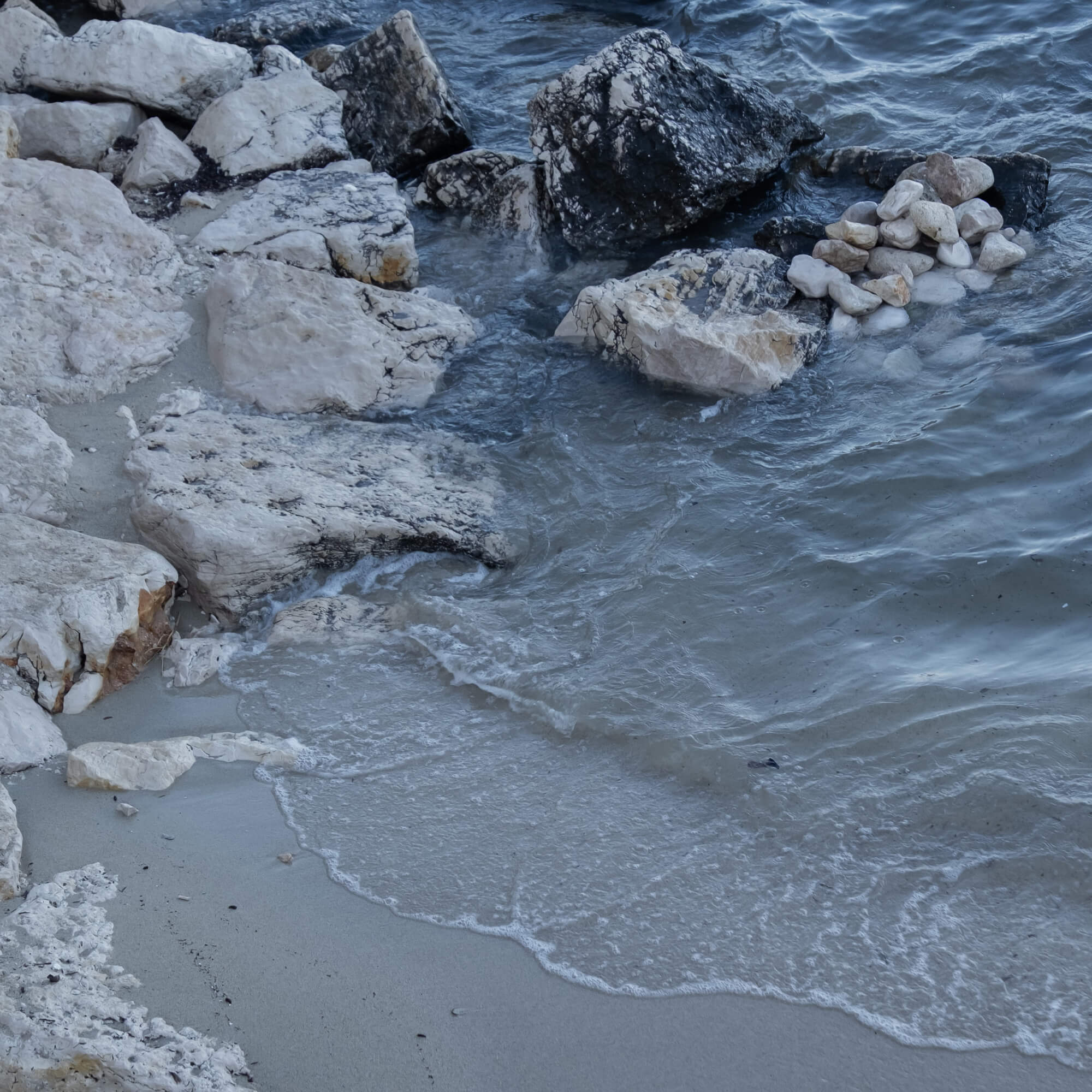 Küstenabschnitt mit hellen und dunklen Steinen und Wellen, die auf den Sand treffen