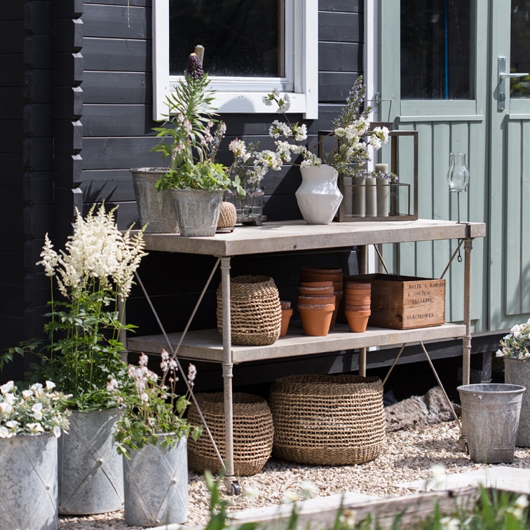 Metall-Gartentisch mit Blumentöpfen, geflochtenen Körben und Blumen vor schwarzer Wand und grüner Tür