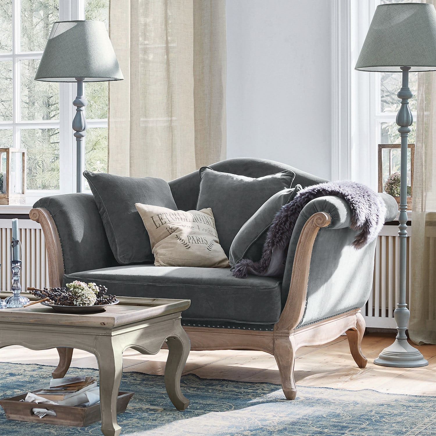 Sessel Sumerset Graues Samtsofa mit Holzrahmen und Kissen in hellem Wohnzimmer mit Stehlampen