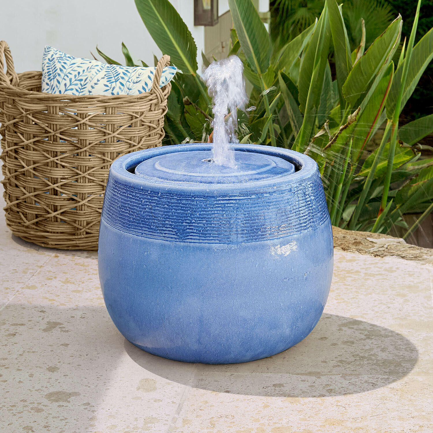 Brunnen Seravia Blaue Keramik-Gartenbrunnen mit spritzendem Wasser vor grünen Pflanzen und Korb aus Weide